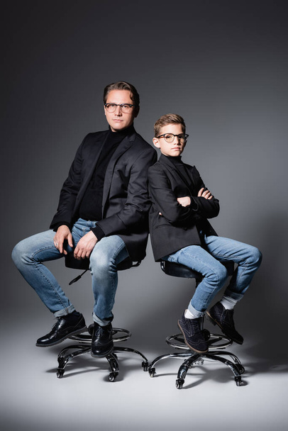 Stylish father and son posing on chairs on grey  - Фото, зображення