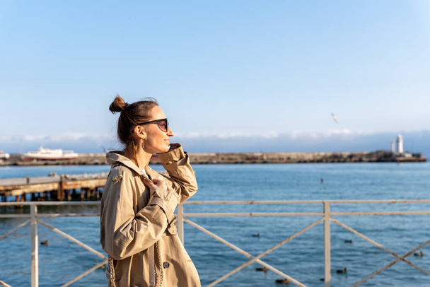 Fiatal felnőtt divatos elegáns gyönyörű kaukázusi boldog mosolygós nő élvezze séta a jaltai tengeri töltés meleg napsütéses napon. Női személy portré viselni farmer biege ballon kabát a városi utcában - Fotó, kép