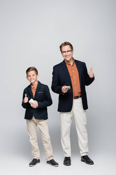 Smiling father and son holding smartphones and showing like on grey background  - Photo, Image