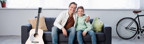 Cheerful father hugging son near acoustic guitar at home, banner  - Фото, зображення