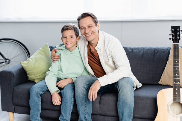 Father hugging preteen boy near acoustic guitar in living room  - Photo, Image