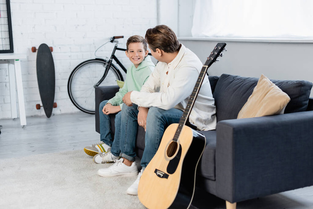 Hymyilevä poika puhuu isälle lähellä akustista kitaraa kotona  - Valokuva, kuva