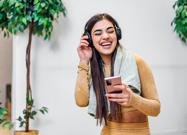 Jonge volwassen glimlachende vrolijke meisje vrouw luisteren naar muziek van smartphone het dragen van een draadloze hoofdtelefoon en dansen tijdens het sporten en het nemen van een pauze van fitness in haar huis - Foto, afbeelding