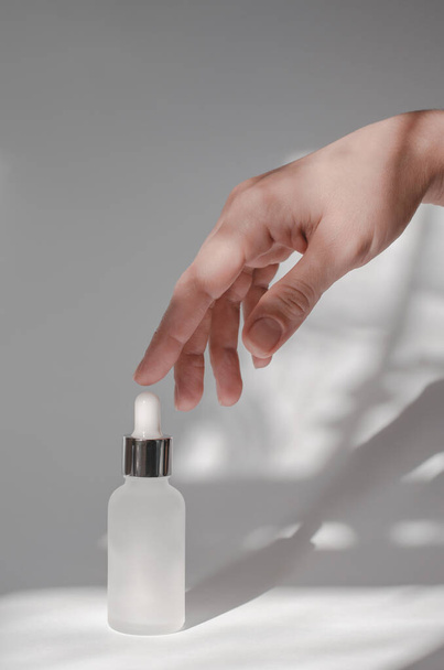 The hand touches a cosmetic bottle with essential oil or serum. White cosmetic bottle and female hand on a white background with shadows - Fotografie, Obrázek