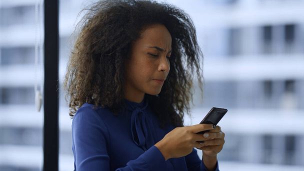 Εστιασμένη αφρικανή αμερικανίδα στέλεχος που ψάχνει κινητό τηλέφωνο στο χώρο εργασίας. - Φωτογραφία, εικόνα