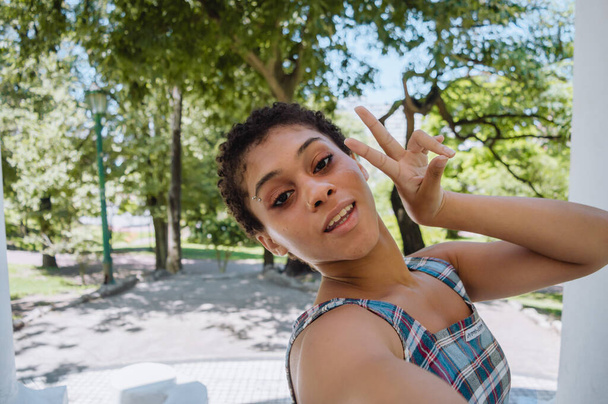 jeune hispanique latino brune femme aux cheveux courts souriant à l'extérieur dans un parc prenant un selfie de paix et d'amour, arbres en arrière-plan avec espace de copie. - Photo, image