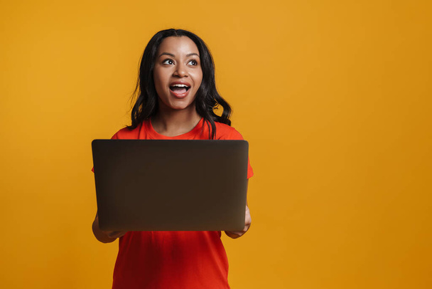 fiatal fekete nő mosolyog, miközben pózol laptop elszigetelt sárga háttér - Fotó, kép