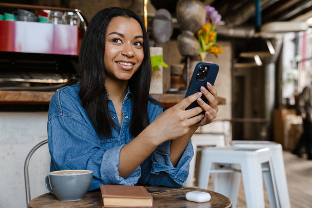 Black smiling woman using cellphone while sitting in cafe - Фото, зображення