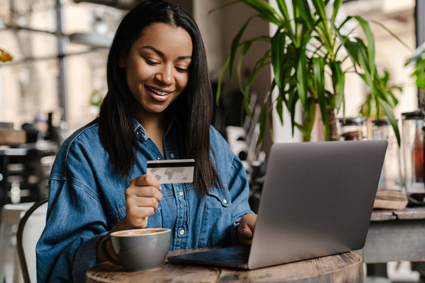 Happy young african woman holding credit card while shopping via laptop computer, sitting at the cafe indoors - Foto, imagen