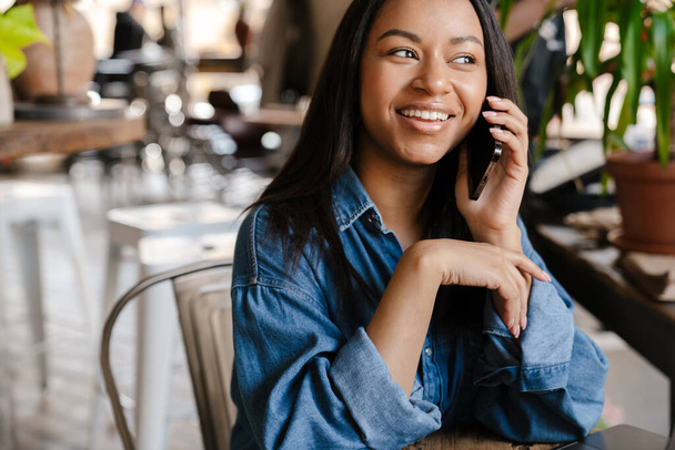 Black smiling woman talking on cellphone while sitting in cafe - Foto, imagen