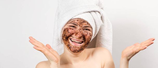 A young woman with a coffee mask on her face and a bath towel on her head smiles and looks into the camera on a white background, a place for text. - Photo, Image