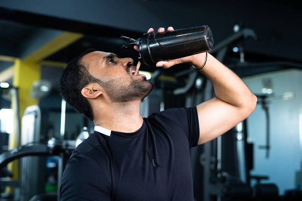 bodybuilder drinking or taking protein powder and mixing with water on bottle by shaking at gym - concpet of muscular gain, fitness training and workout - Foto, Imagem