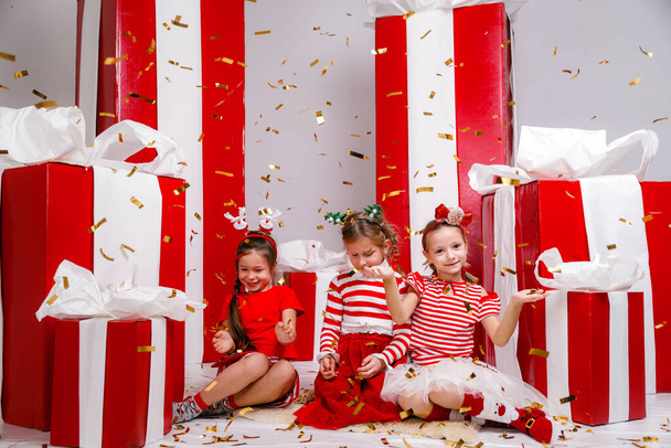 grappige kleine meisjes vrienden in kerstkostuums tegen de achtergrond van grote rode cadeautjes. idee en concept van een gelukkige vakantie, nieuw jaar, vriendschap en jeugd. - Foto, afbeelding