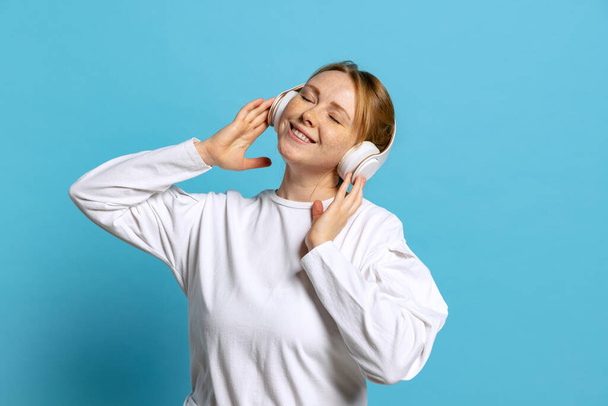 Dreaming young beautiful girl wearing white pullover listening to music isolated on blue background. Concept of emotions, facial expression, youth, aspiration, sales - Фото, изображение