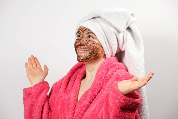 Una hermosa mujer con un exfoliante de café en la cara en un albornoz con una toalla en la cabeza sobre un fondo blanco hace que los procedimientos de cuidado de la piel del spa - Foto, imagen