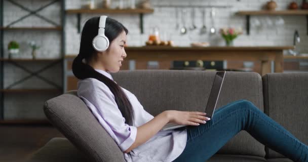 Asian business woman with headphones video chatting with laptop on couch at home. Female student talking and looking at laptop screen while sitting on couch at home living room. Webcam webinar concept - Footage, Video