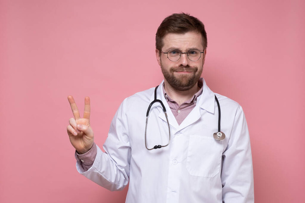 聴診器付きの疲れた男性医師は勝利の象徴を示しています。2本の指でジェスチャー。ピンクの背景. - 写真・画像