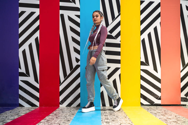 Jonge vrouw staande kijkend naar camera tegen muur met kleuren en gestreepte ontwerpen - Foto, afbeelding