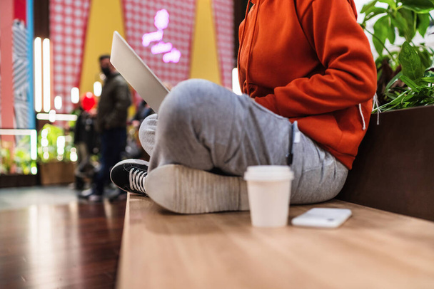 Vágott kilátás egy felismerhetetlen fiatal nő ül a padon keresztbe tett lábakkal használja a laptopját egy bevásárlóközpontban - Fotó, kép