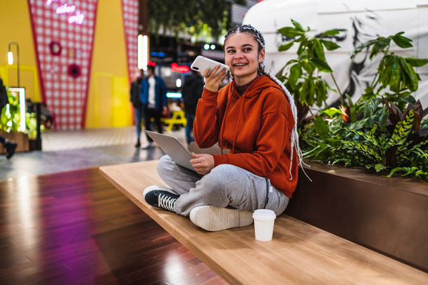 Eine junge Frau sitzt mit ihrem Laptop auf einer Bank und nimmt eine Audiobotschaft in einem Einkaufszentrum auf - Foto, Bild