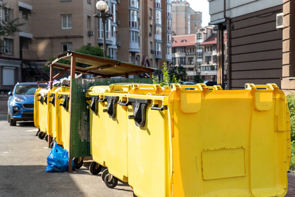 都心部や郊外の住宅街の建物の近くに黒いプラスチックごみ袋でいっぱいの多くの大きなプラスチック製の黄色のゴミ箱の行。リサイクルできない分別ガベージコレクション - 写真・画像