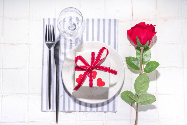  San Valentino tavola apparecchiata con piatto, confezione regalo con nastro rosso festivo, bicchiere di vino, forchetta e coltello, rose rosse bouquet fiori bianco piastrellato tavolo flatlay copia spazio - Foto, immagini