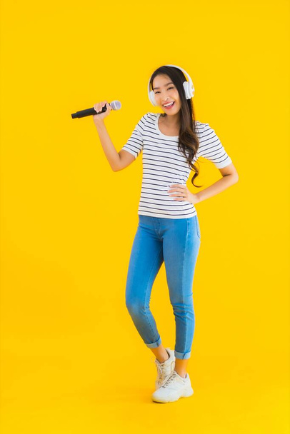 Retrato hermosa joven asiática mujer cantando con micrófono y auriculares en amarillo aislado fondo - Foto, imagen