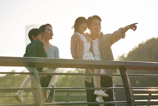 Asijská rodina se dvěma dětmi stojící na pěší most při pohledu na pohled v městském parku za soumraku - Fotografie, Obrázek