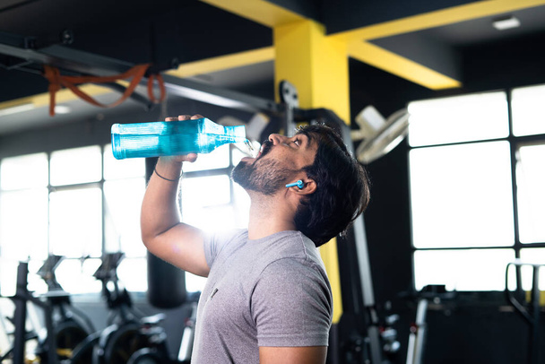 Sed indio joven musclebuilder beber agua después del entrenamiento en el gimnasio - concepto de relajación, estilos de vida saludables y entrenamiento duro o sesión de entrenamiento de fitness. - Foto, imagen