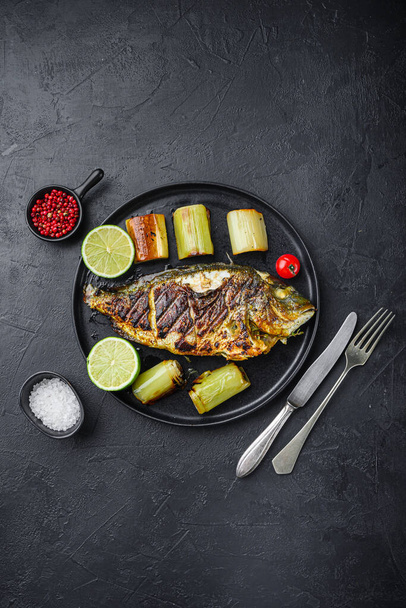 Gegrillte Dorade oder Dorade roher Fisch auf schwarzem Teller mit geschmortem Lauch auf schwarzem Hintergrund, Ansicht von oben - Foto, Bild