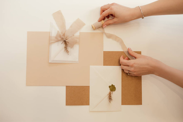 Обрізана рука жінки, персиковий композиційний папір, запрошені конверти, стрічки, віск, вирізані роботи ручної роботи. Вид зверху
 - Фото, зображення