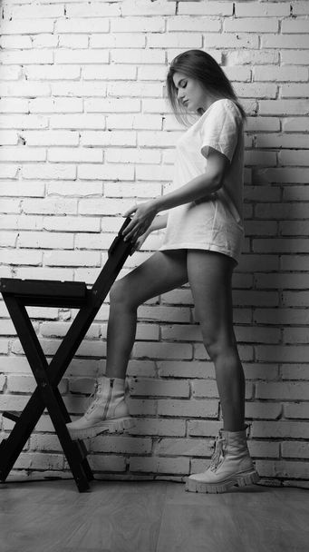 Fekete-fehér fotó fiatal gyönyörű nő hosszú haj visel t-shirt pózol közelében szék stúdióban.   - Fotó, kép