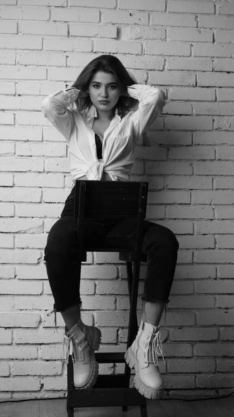 Photo noir et blanc de jeune belle femme sensuelle et sexy aux cheveux longs portant une chemise blanche posant contre un mur de briques blanches en studio.   - Photo, image