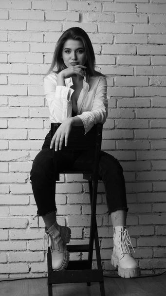Foto en blanco y negro de una joven hermosa mujer sensual y sexy con el pelo largo usando una camisa blanca posando contra una pared de ladrillo blanco en el estudio.   - Foto, imagen