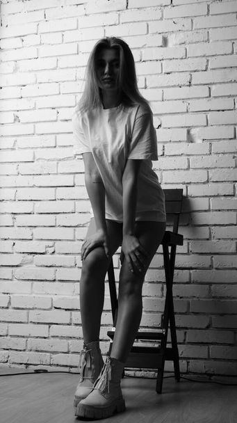 Black and white photo of young beautiful woman with long hair wearing in t-shirt posing with chair in studio.   - Fotoğraf, Görsel