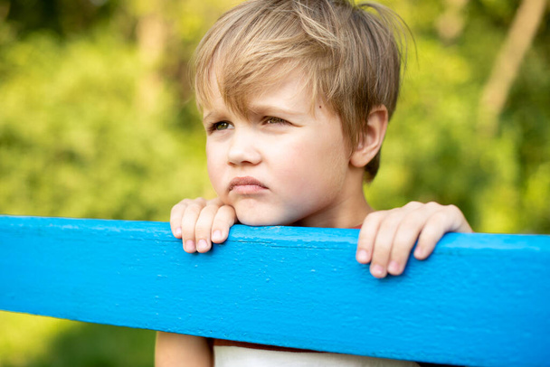 Portrait d'un beau garçon sur un fond de parc verdoyant près de la clôture bleue. Le gars regarde attentivement sur le côté - Photo, image