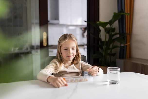 Little Caucasian Girl in pajamas taking natural vitamin omega-3 from plastic box on kitchen at home early morning. Adorable child Takes supplements. - 写真・画像