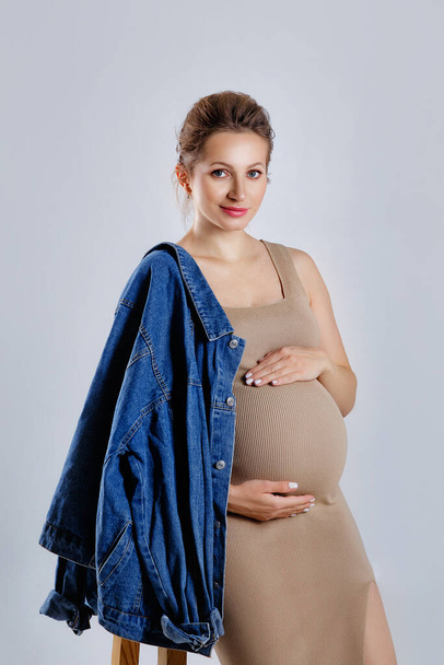 egy gyönyörű terhes szőke egy bézs ruha és farmer, álló szürke stúdió háttér, a terhesség divat, üzletasszony anya - Fotó, kép