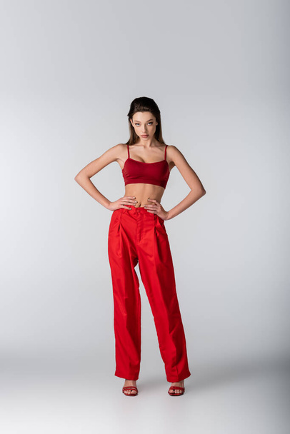 full length of pretty model in red outfit posing with hands on hips on grey - Φωτογραφία, εικόνα