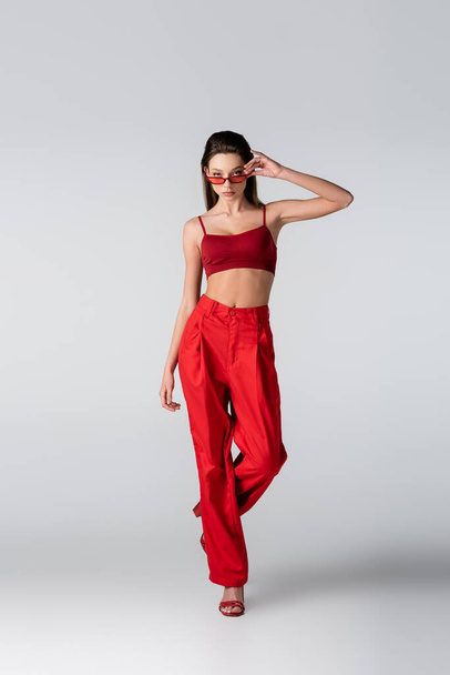 full length of young model in red outfit adjusting sunglasses on grey - Фото, изображение