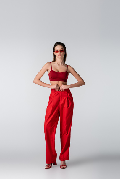 full length of young woman in sunglasses and red outfit posing on grey - 写真・画像