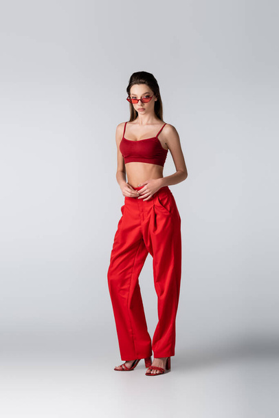 full length of young model in sunglasses and red outfit standing on grey - Фото, изображение