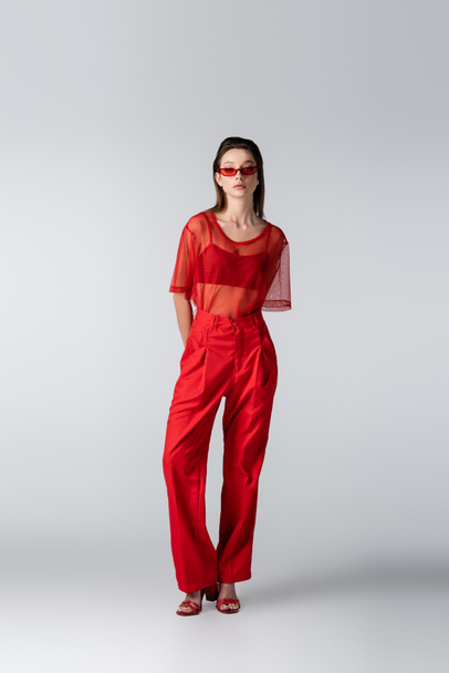 full length of young model in sunglasses and red trendy outfit posing on grey - Foto, Imagem