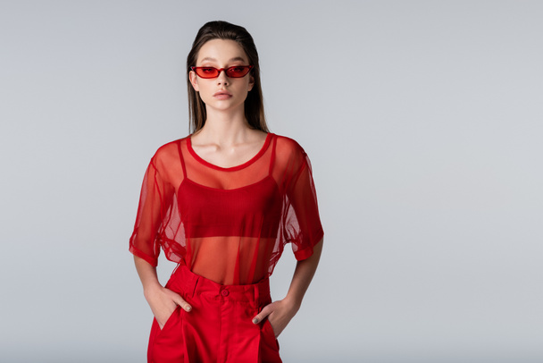 young model in red trendy outfit and sunglasses posing with hands in pockets isolated on grey - Фото, изображение