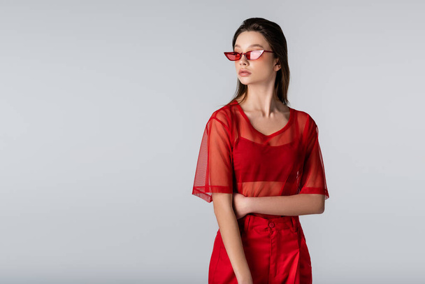 young model in trendy outfit and red sunglasses posing isolated on grey - Foto, imagen