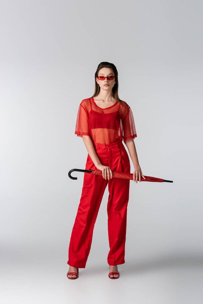 full length of young woman in red trendy outfit and sunglasses holding umbrella on grey - Foto, Imagen