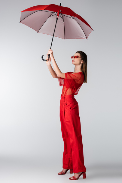 longitud completa de modelo joven en traje de moda rojo y gafas de sol con paraguas por encima de la cabeza en gris - Foto, Imagen
