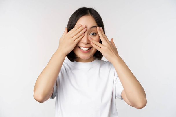 Портрет азиатки, закрывающей глаза, ожидающей сюрприза с завязанными глазами, улыбающейся и смотрящей в камеру, стоящей на белом фоне - Фото, изображение
