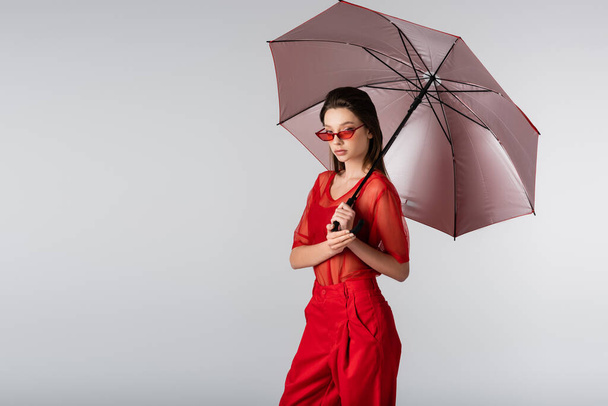 young woman in red outfit and sunglasses standing under umbrella isolated on grey - Foto, imagen