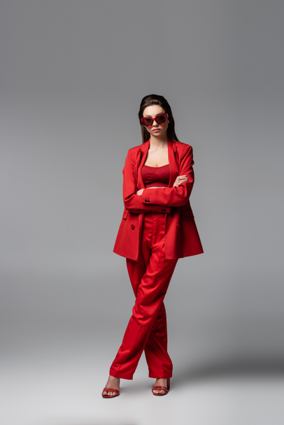 full length of young woman in trendy red suit and sunglasses standing with crossed arms on dark grey - Photo, Image